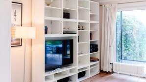 A modern living room with a flat-screen TV, white shelving, a floor lamp, and a view through a window. - Holiday Home 'Seeblick' with Sea View, Private Terrace and Wi-Fi (Langballig)