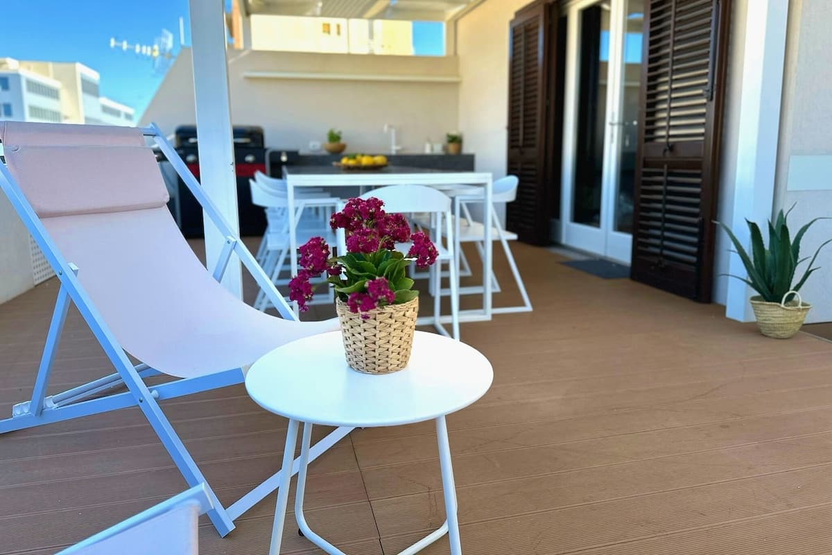 A balcony with a lounge chair, a small table with a woven basket of flowers, and a potted plant.