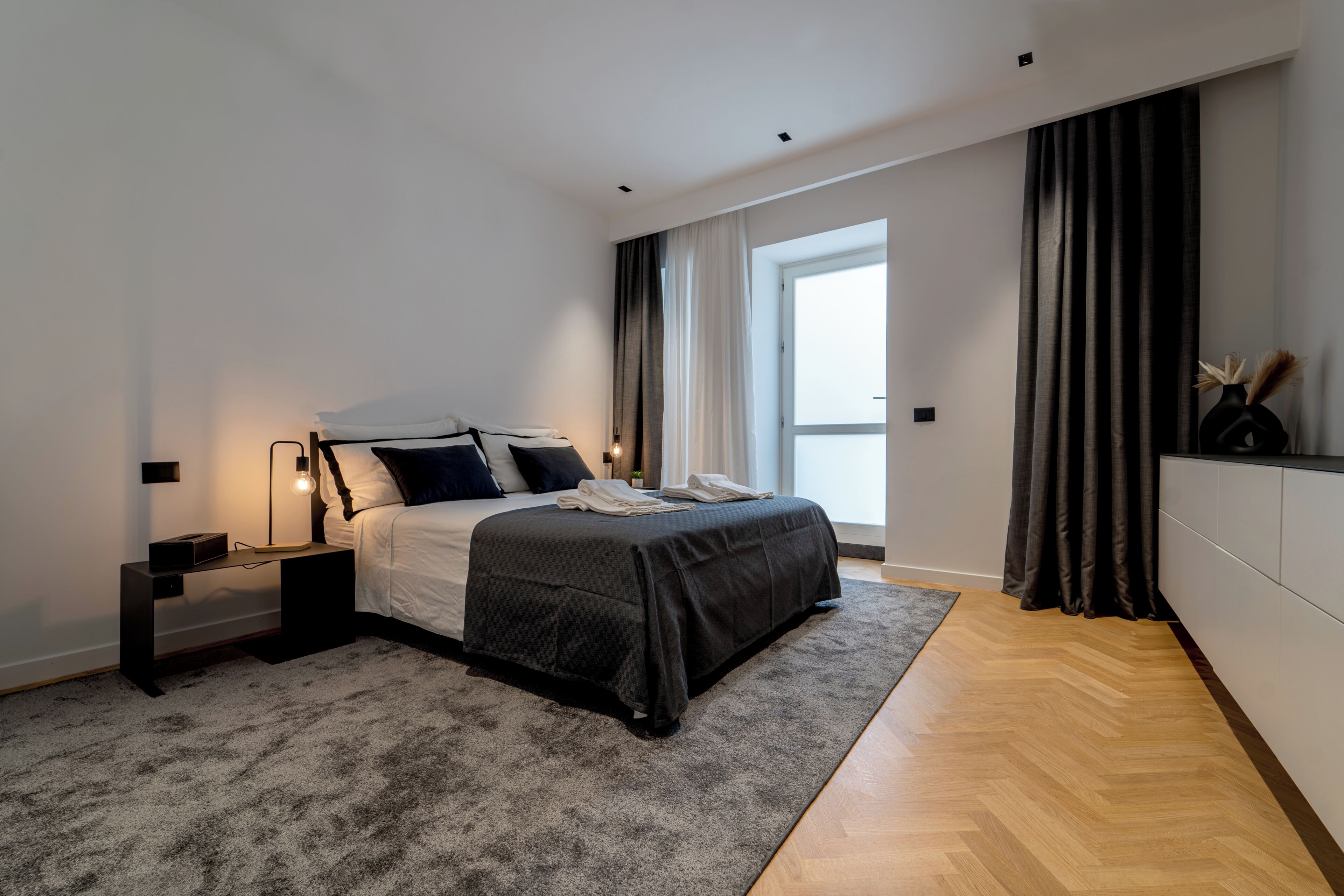 A modern bedroom with a bed, bedside tables, a lamp, and a view of the outdoors.