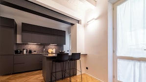 A modern kitchen with dark cabinetry, a herringbone wood floor, and a large window with sheer curtains. - Gonia Residence (Trapani)