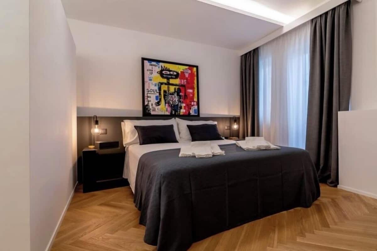 A modern hotel room with a large bed, a nightstand, a wall-mounted artwork, and a window with curtains.