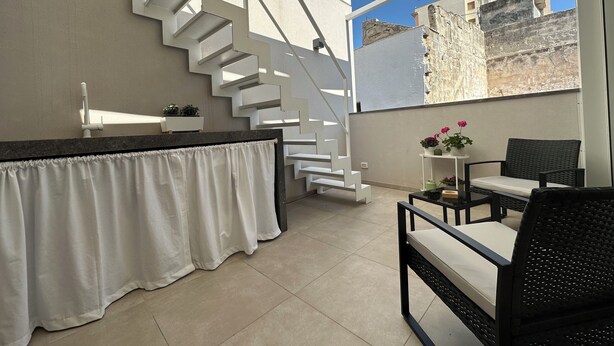A modern kitchen with a white staircase, a bar area with a white countertop, and a view of the cityscape through large windows. - Gonia Residence (Trapani)