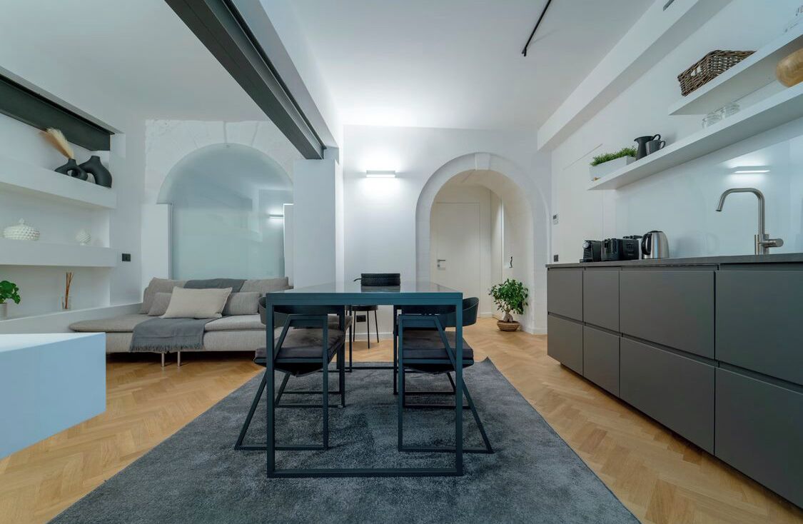 Un saló modern amb un sofà, una taula de dinar amb cadires, una cuina amb armaris i una escala cap a una planta superior.