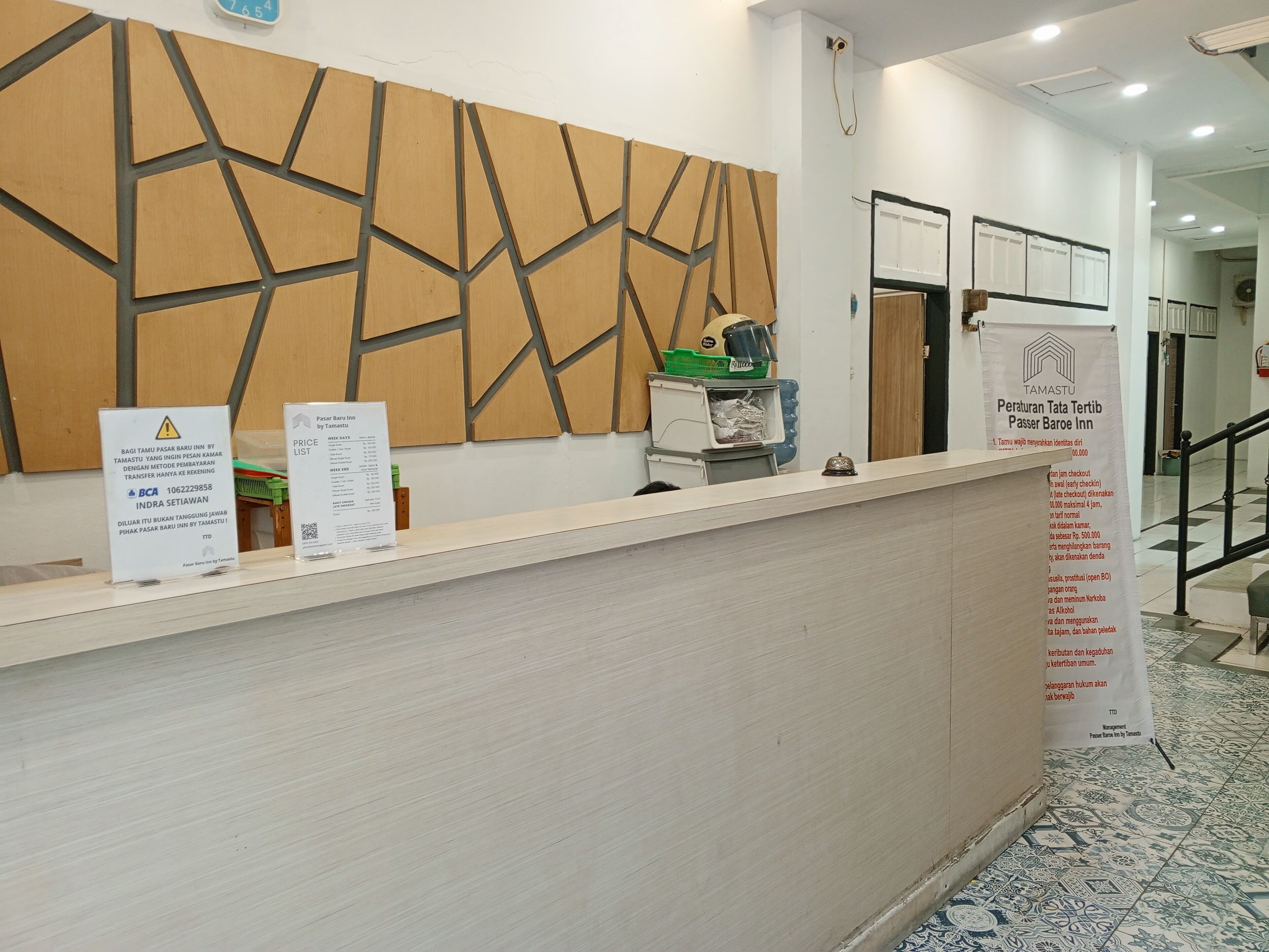 Un comptoir d’accueil d’hôtel au design moderne, un panneau indiquant les tarifs des chambres et un tableau d’affichage mural contenant des informations.