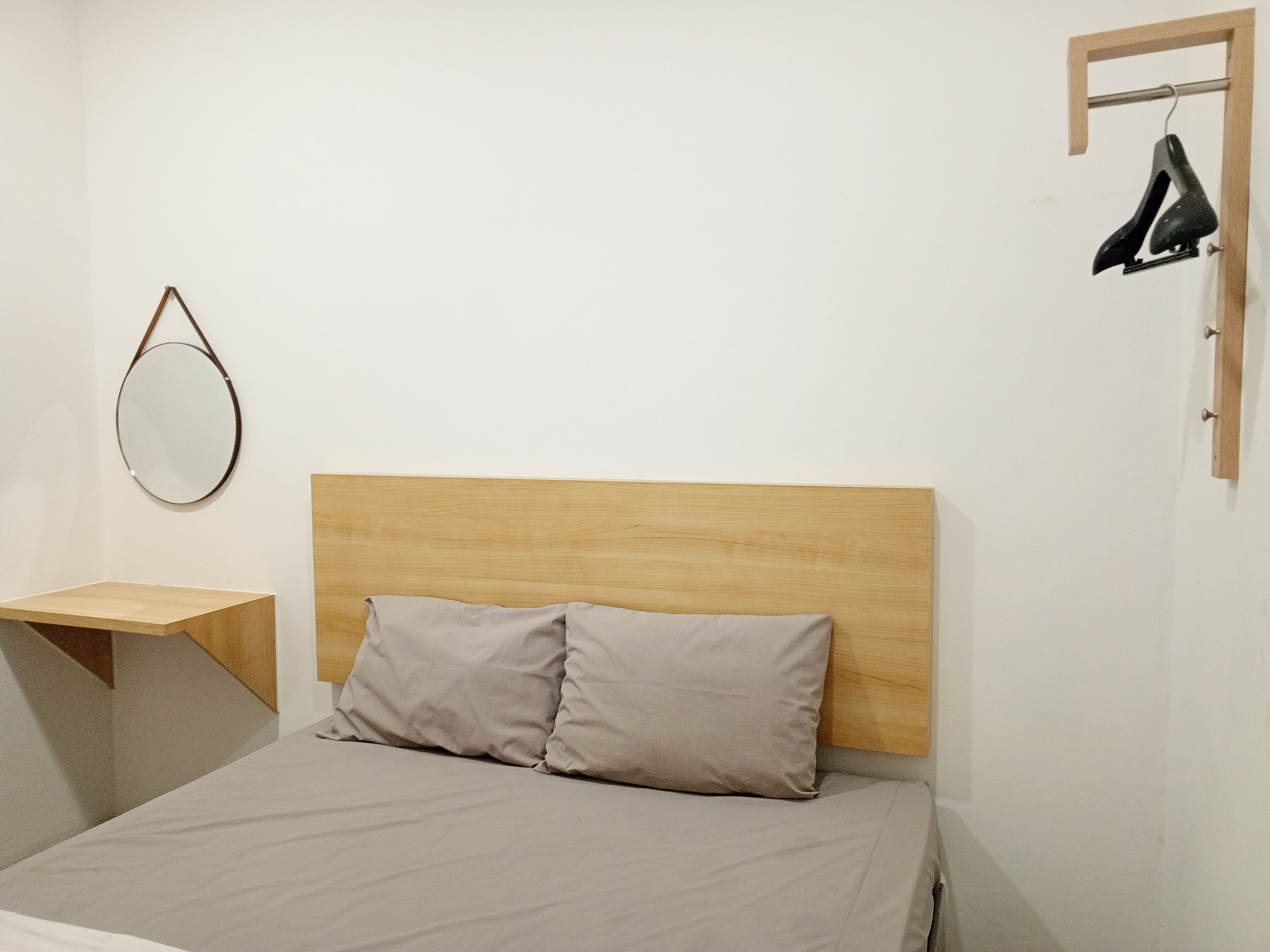 A bedroom with a wooden headboard, a bed with two pillows, a small wooden nightstand, a round mirror, and a shoe rack with a single shoe.