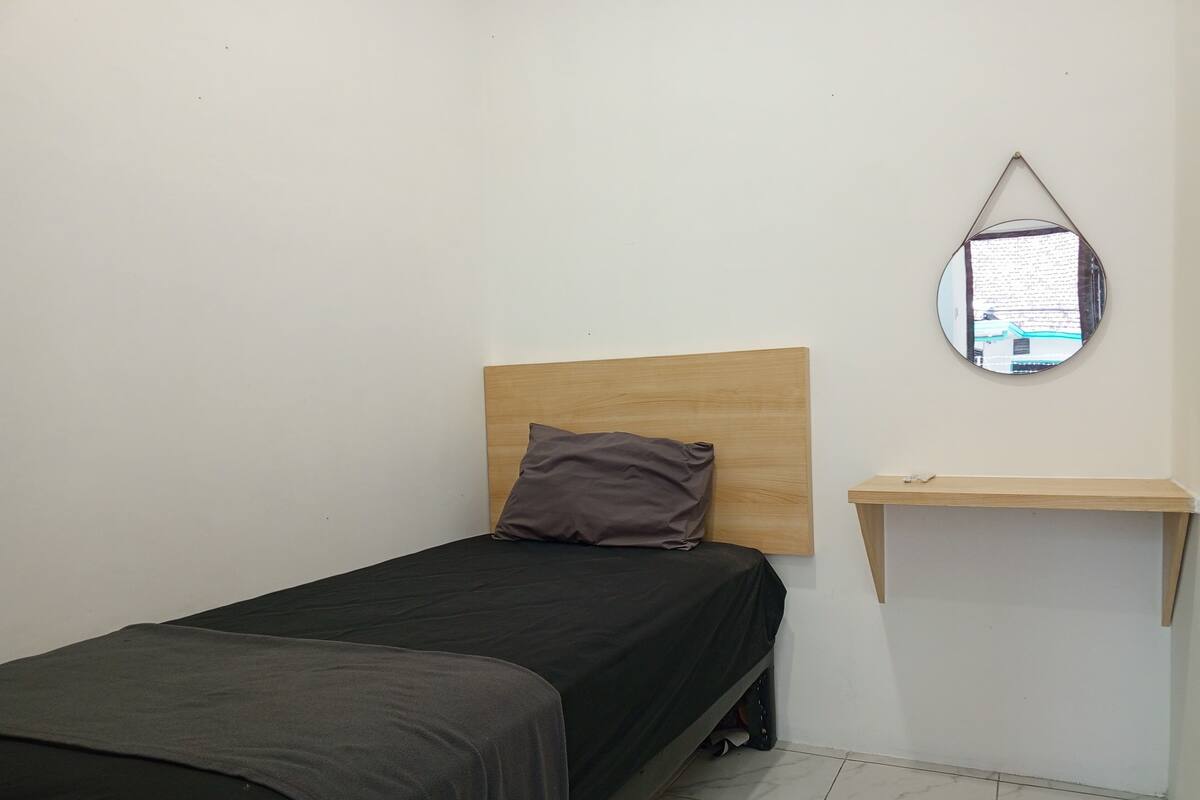 Una habitación pequeña con una cama individual, cabecera de madera, una almohada, un estante de madera pequeño y un espejo en la pared.