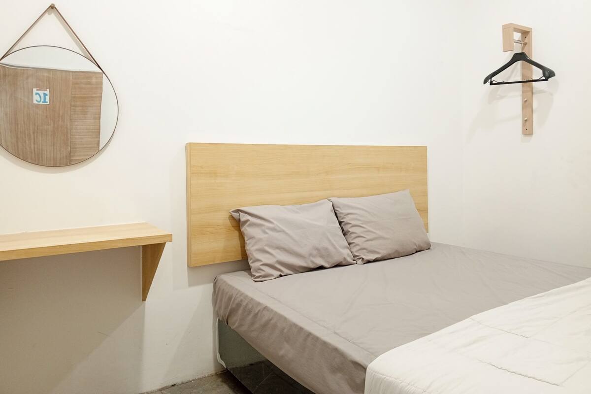Un dormitorio con cabecera de madera, cama con colchón y almohadas color beige, una mesita de noche de madera y un perchero de madera fijado a la pared.