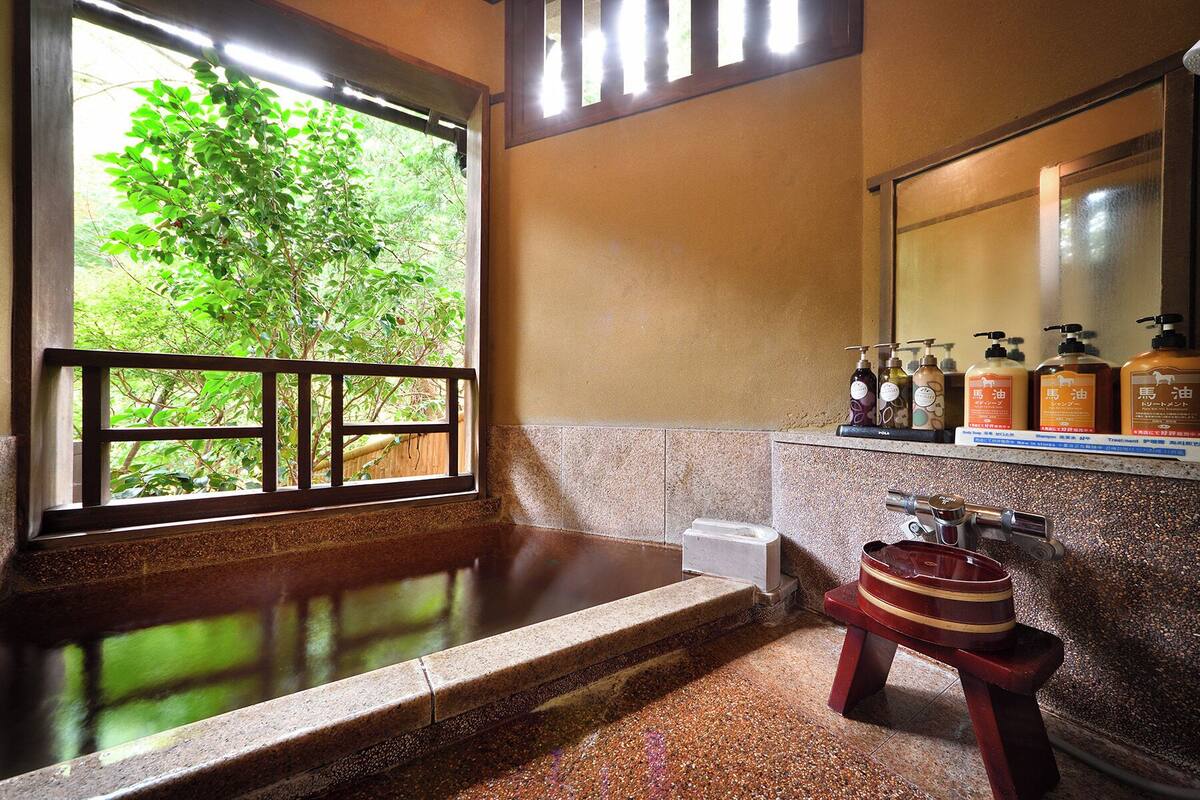 Un baño con un ventanal grande, una bañera, un taburete de madera y artículos de aseo en la encimera.