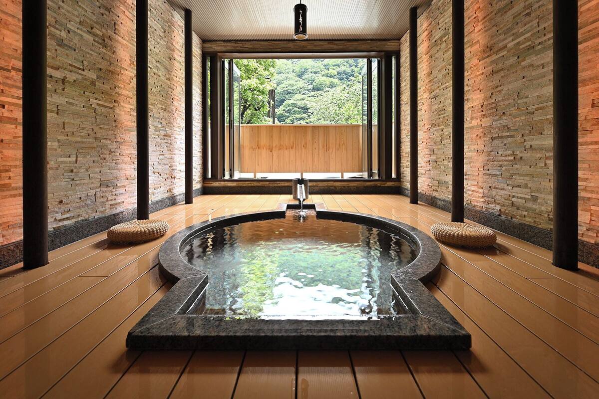 Un área de spa con un jacuzzi central, suelo de madera, paredes de ladrillo y amplios ventanales con vistas a zonas verdes.