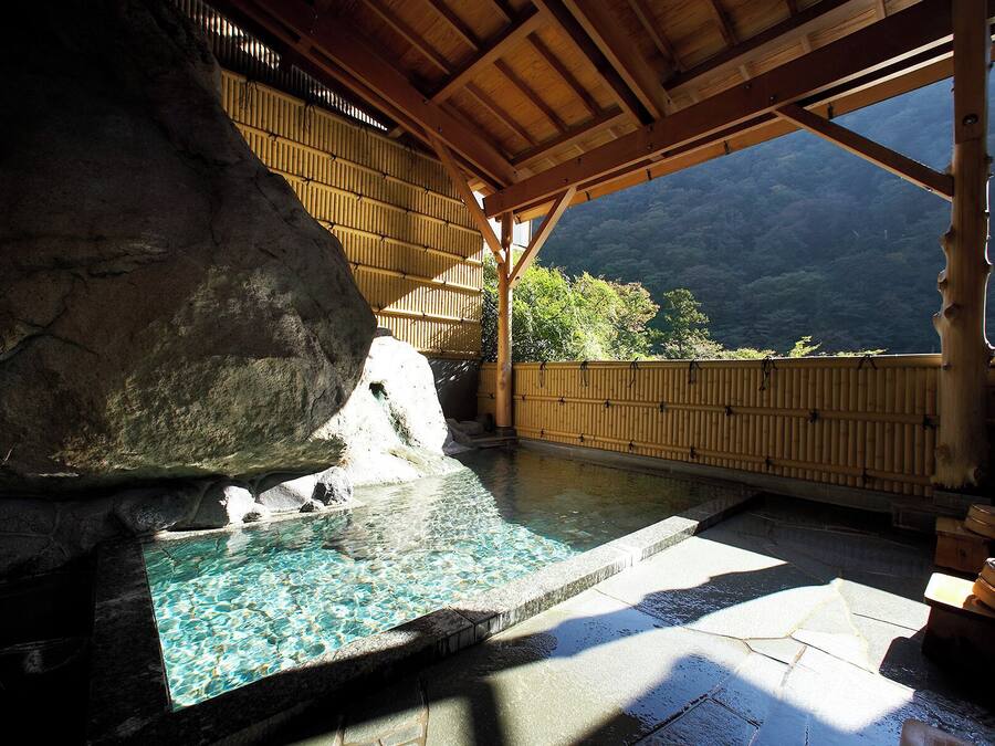 A hot spring bath area with a wooden roof, stone walls, and a view of mountains.