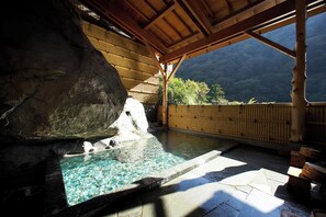 Un espace de baignade dans une source thermale, doté d’un toit en bois, de murs en pierre et offrant une vue sur les montagnes.