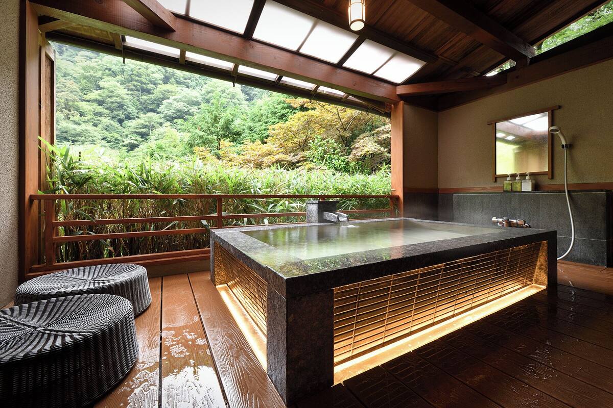 Un onsen japonés tradicional con una terraza de madera, una bañera de piedra caliente y vistas de abundante vegetación.