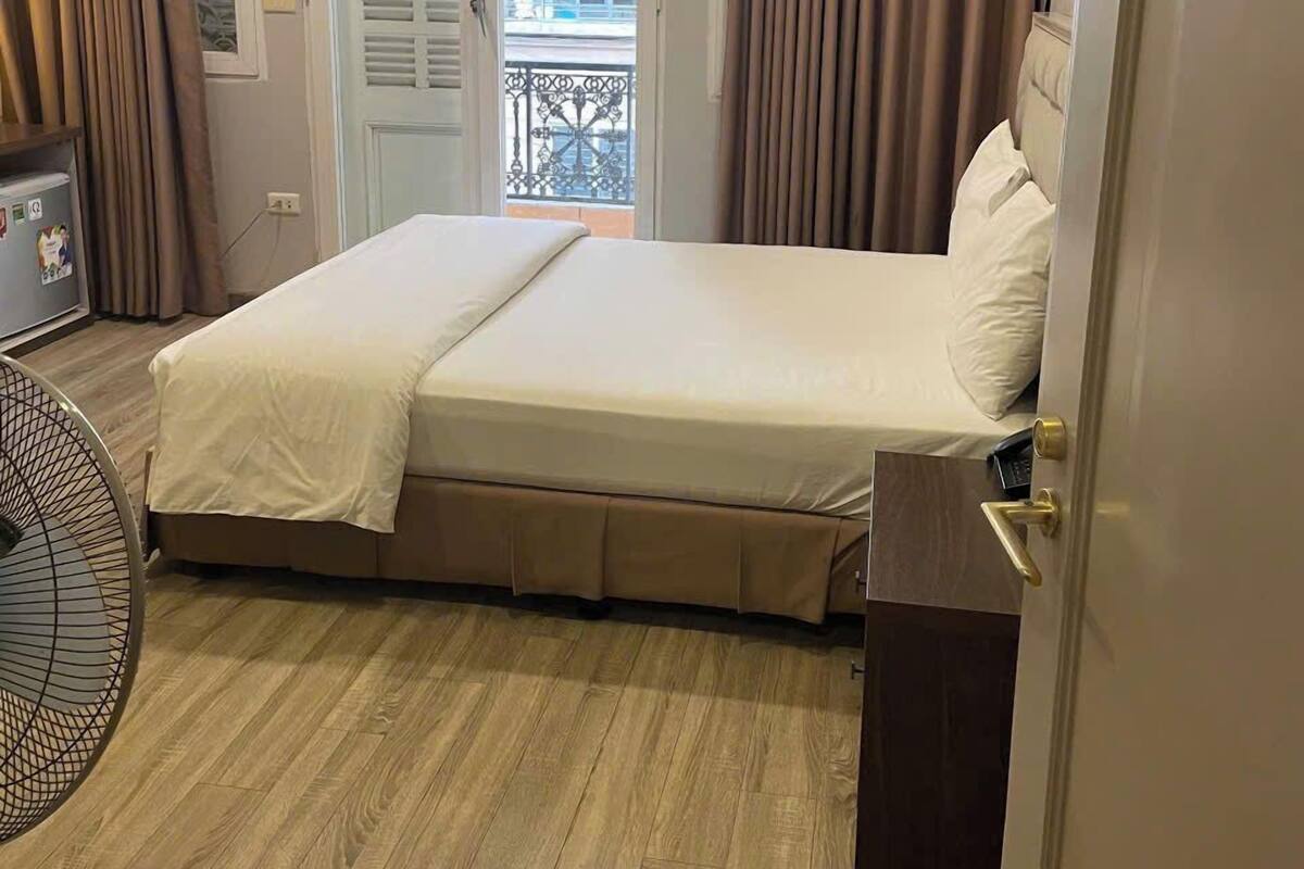 Habitación de hotel con cama, ventilador, una mesita y vistas al balcón exterior.