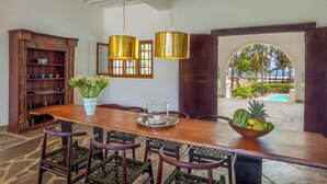 A dining area with a wooden table, chairs, a fruit bowl, a vase with flowers, and a bookshelf. - Marula House 3BD, Diani Beach (Diani Beach)