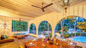 A dining area with a wooden table set for a meal, a ceiling fan, and a view of a swimming pool and greenery. - Marula House 3BD, Diani Beach (Diani Beach)