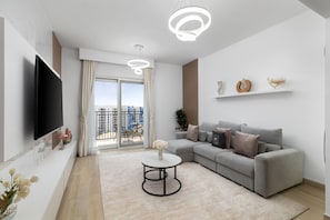 Ein modernes Wohnzimmer mit grauem Sofa, einem flachbildfernseher an der Wand, einem runden Couchtisch mit Blumenvase und einem Balkon mit Blick auf die Stadt.