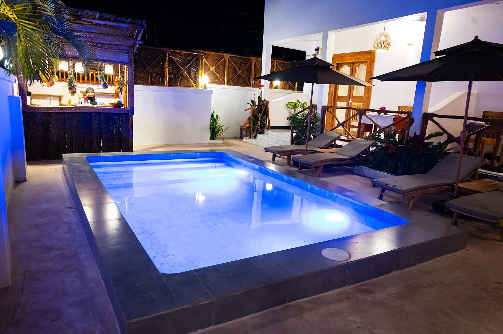 Una piscina con sillones de descanso y sombrillas, rodeada por una zona de patio con plantas y un bar.