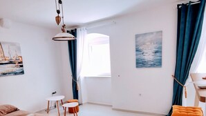 Une pièce moderne et bien éclairée, avec un lit, une petite table, une chaise et un canapé. Deux tableaux ornent les murs : l’un représente un voilier et l’autre, un paysage marin.