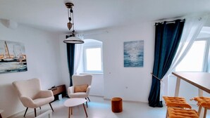 Un salon moderne avec un canapé blanc, un fauteuil blanc, une petite table ronde et un tabouret en bois avec des coussins orange. Un tableau représentant des voiliers est accroché au mur, et un autre tableau représentant de l’eau est accroché au mur opposé.