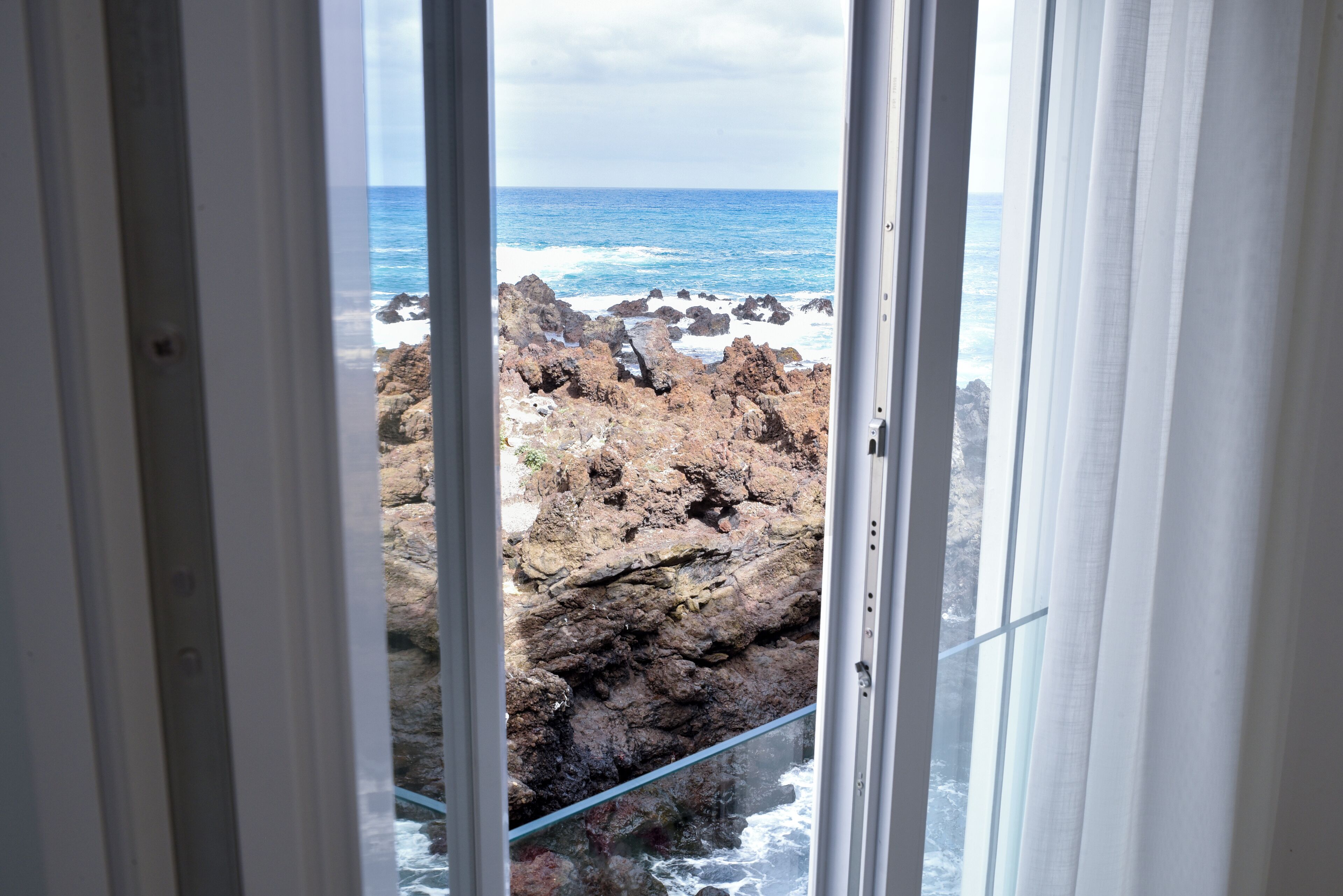 Vista a través de una ventana con cortinas blancas, mostrando terreno rocoso y el océano.