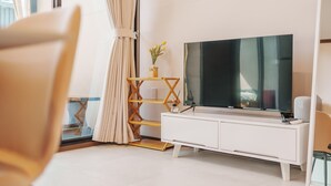 Ein modernes Wohnzimmer mit einem großen Flachbildfernseher auf einem weißen TV-Schrank, einem Holzregal mit einer Pflanze und einem Vorhang.