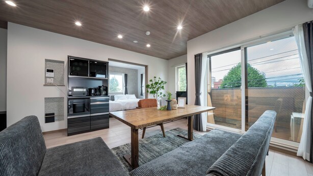 A modern living room with a wooden dining table, grey sofas, and a kitchen area with built-in appliances. - MKS201 (Asahikawa)