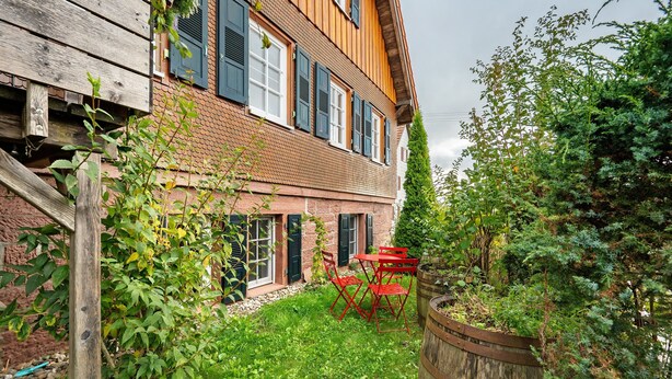 A two-story building with a brick facade, a wooden deck, and a red outdoor table and chairs. - Apartment 'Malia' with Shared Terrace, Shared Garden and Wi-Fi (Neubulach)