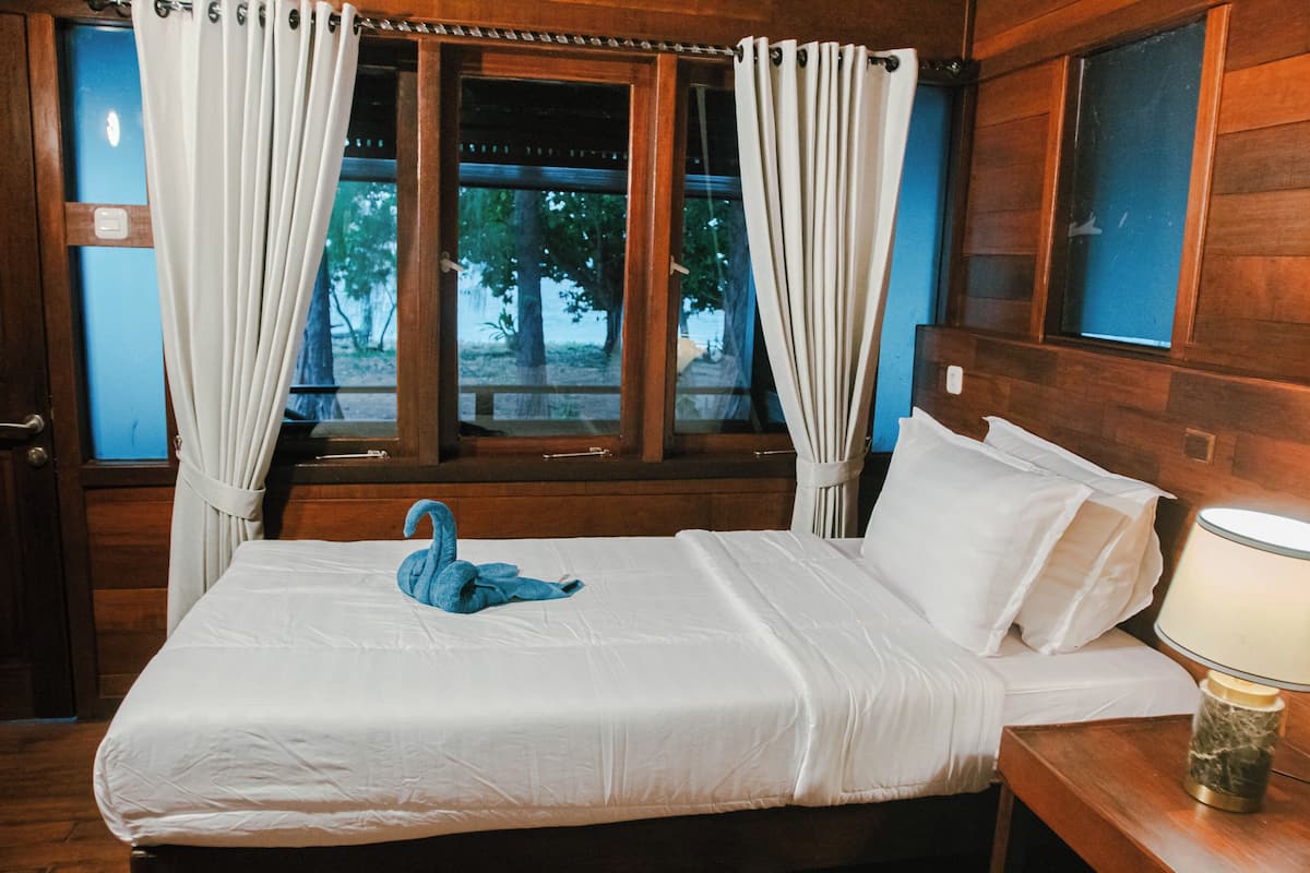 A wooden cabin room with a bed, white curtains, a window with a view of trees, and a lamp on a bedside table.