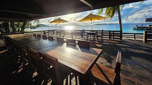 An outdoor dining area with wooden tables and chairs, umbrellas, and a view of the ocean. - ANDAU DIVE RESORT - RAJA AMPAT (Miosandau Kecil Island)