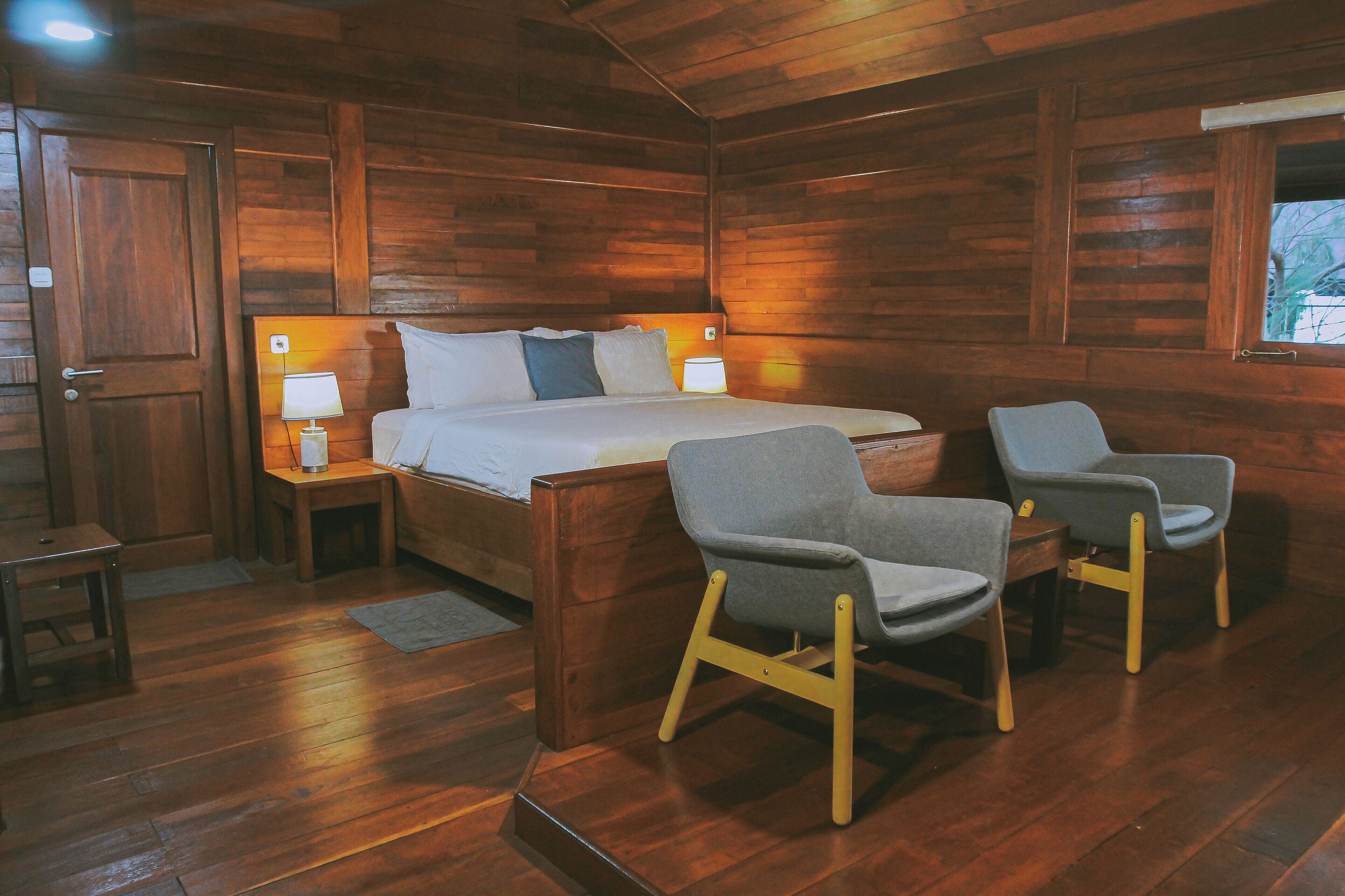 Habitación de cabaña de madera con cama, dos sillas, una mesita de noche y una ventana.
