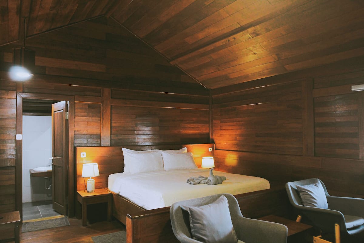 A wooden cabin-style room with a bed, two bedside tables with lamps, a chair, and a small table.