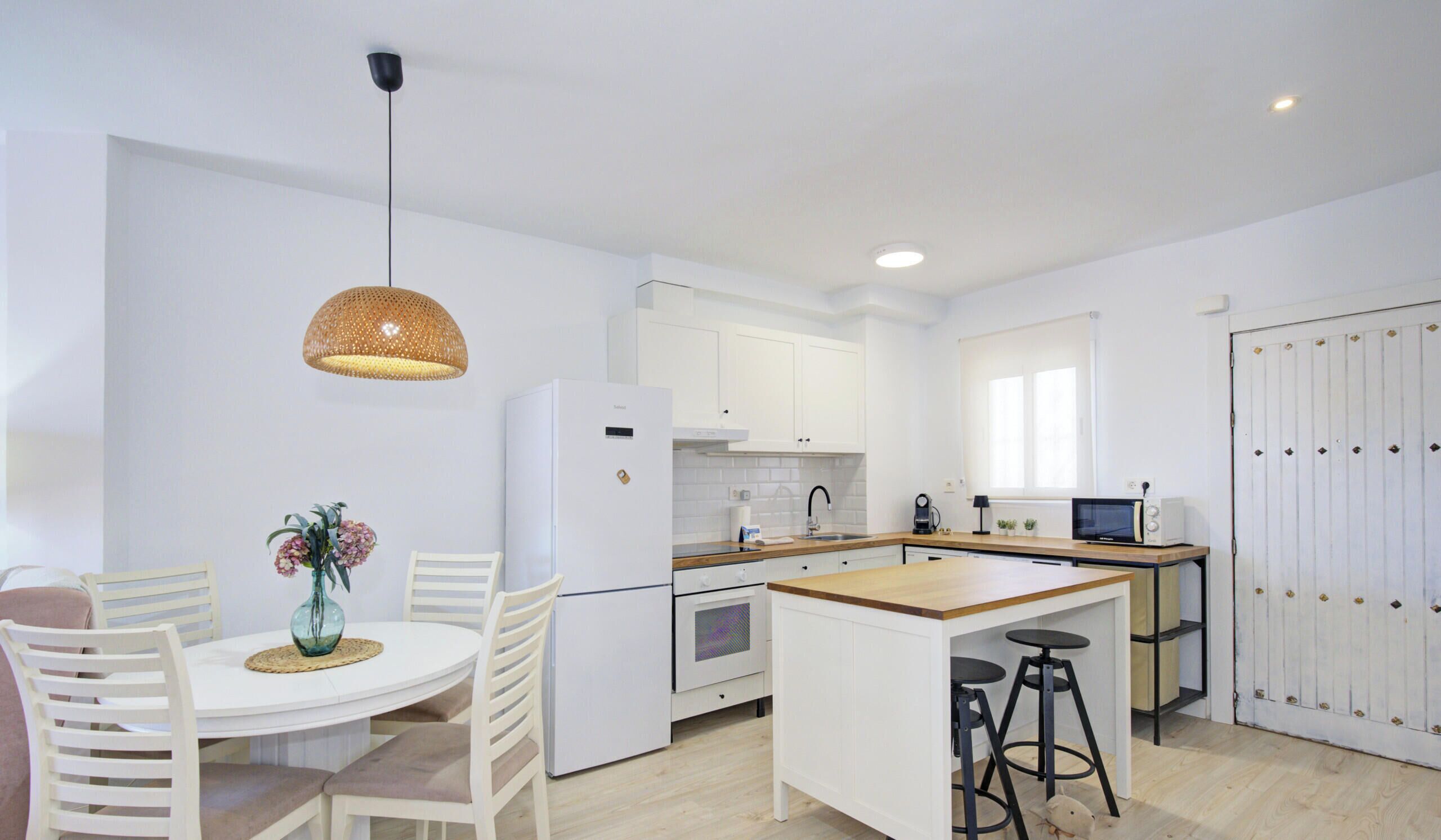Een moderne keuken met witte kastjes, een centraal eiland, een eettafel met stoelen, een hanglamp en een raam dat natuurlijk licht binnenlaat.