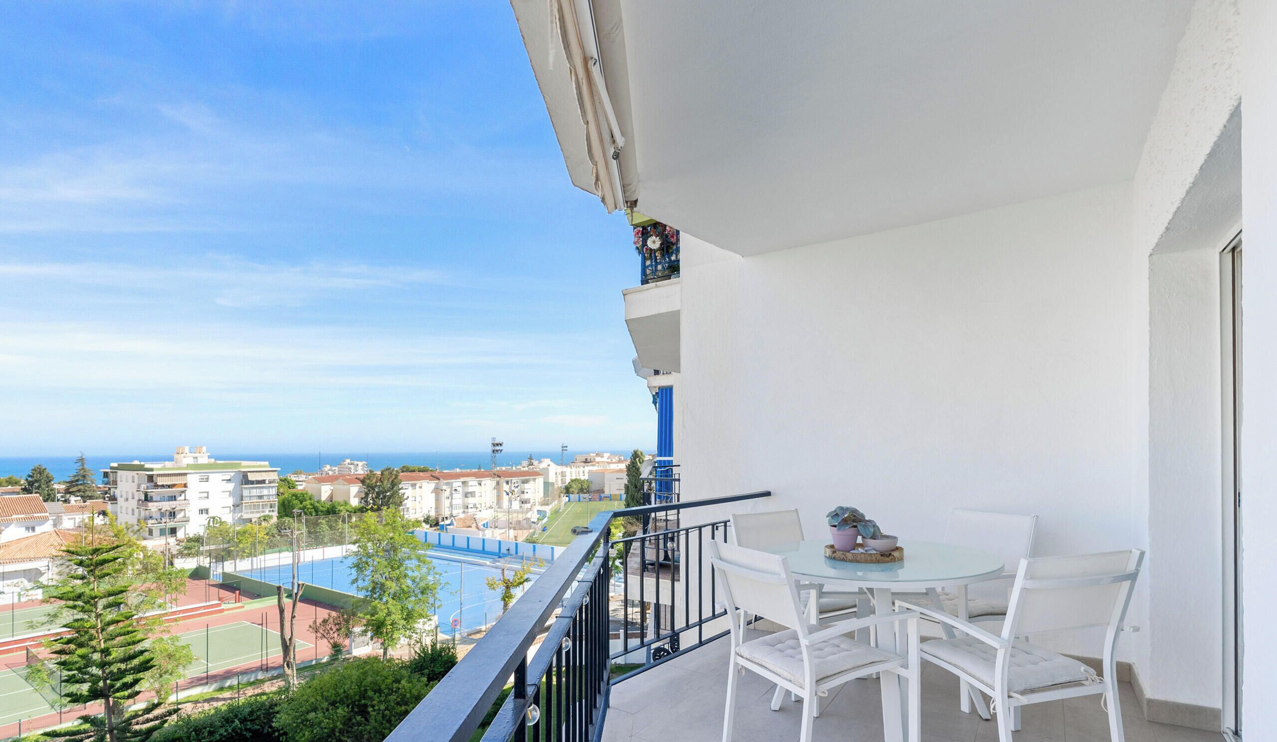 A balcony with white furniture, a view of a tennis court, and buildings in the background.