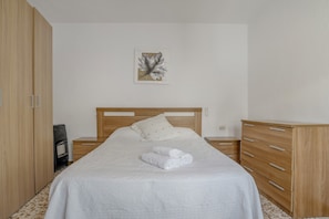A neatly made bed with a white comforter and rolled towels, a wooden headboard, a small bedside table with a lamp, and a wooden dresser with drawers.