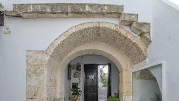 Un arco de entrada conduce a un cuarto con una puerta negra, una pared blanca y una pequeña planta en maceta.