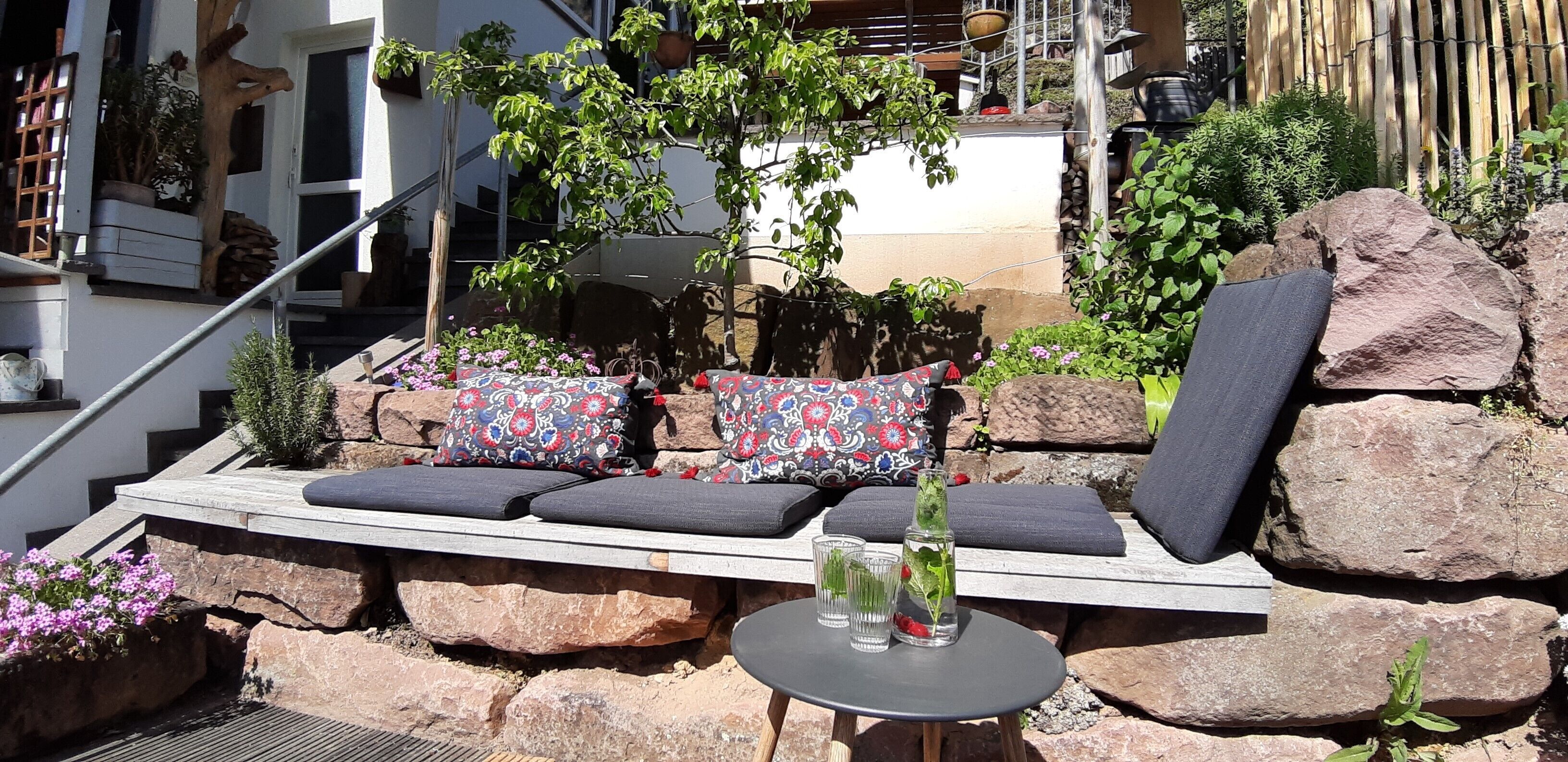 Eine Steinsitzbank mit Kissen, ein kleiner Tisch mit Gläsern und ein Blumentopf.