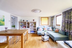 A living room with a wooden dining table, a sofa with cushions, a bookshelf, and a window with curtains.