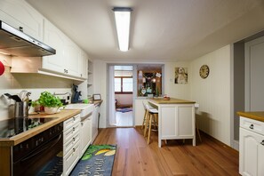A kitchen with white cabinets, a stove, a sink, and a wooden countertop. There is a window with blinds, a door leading to another room, and a clock on the wall.