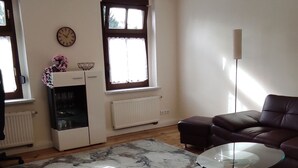 A living room with a wooden floor, a glass coffee table, a brown leather sofa, a modern chandelier, and a large window with blinds. - Apartment Wernerstr in the center - 3 bedrooms (Cottbus)