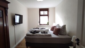 A narrow room with a bed, a TV mounted on the wall, a window with curtains, and a wooden door. - Apartment Wernerstr in the center - 3 bedrooms (Cottbus)