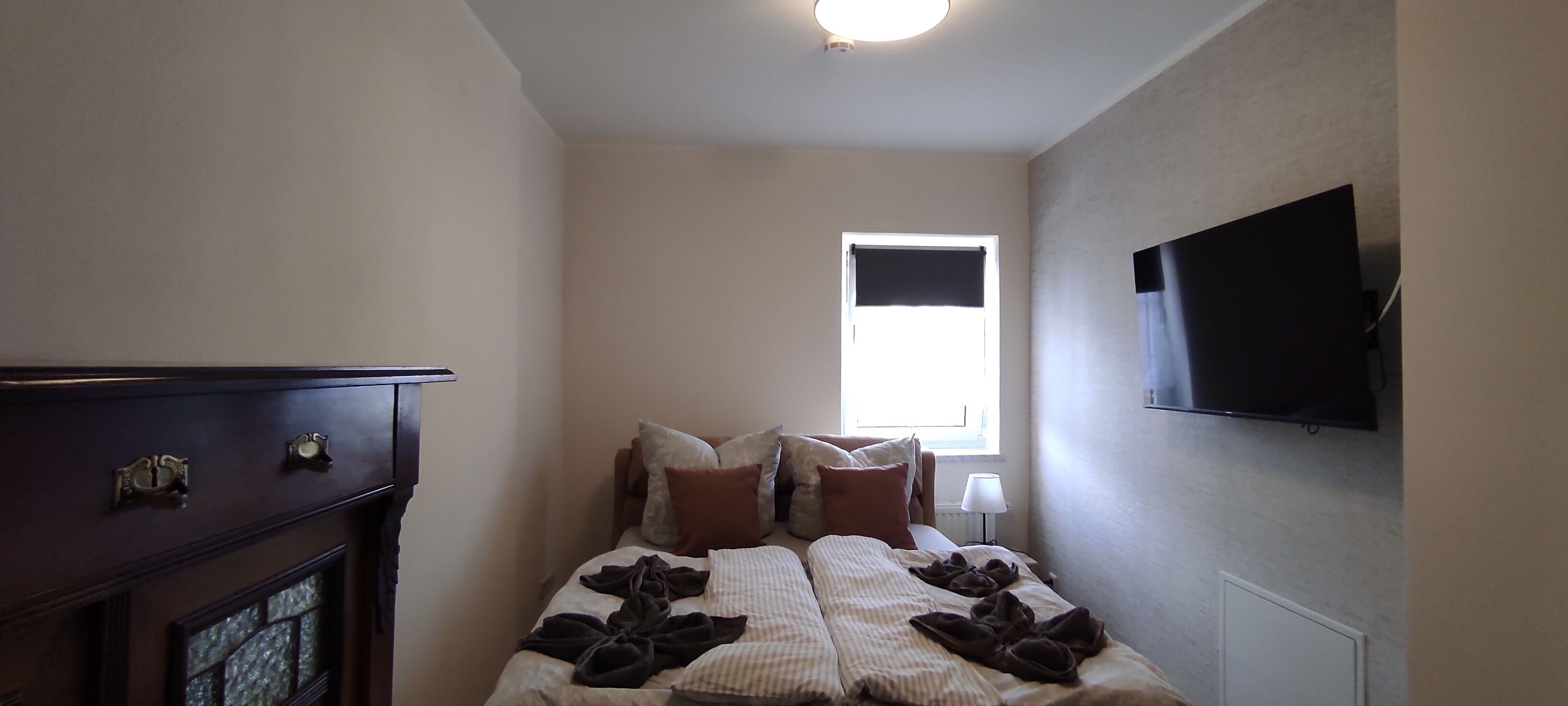 Habitación de hotel estrecha con cama, cómoda, televisor montado en la pared y ventana con vistas.