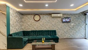 A living room with a green sectional sofa, a glass coffee table with a plant, a wall clock, and a TV mounted on the wall. - Emerald Suites By Classio (Bengaluru)