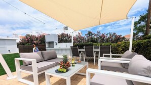 Un patio al aire libre con mobiliario blanco, una mesa con fruta y una botella, y un toldo grande.