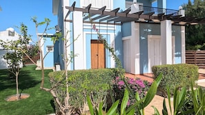Un edificio moderno de dos pisos con fachada azul y blanca, una puerta de madera y una pérgola con plantas trepadoras.
