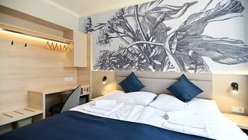 A modern hotel room with a large bed, two pillows, a desk with a chair, a hanging coat rack, and a wall-mounted light.