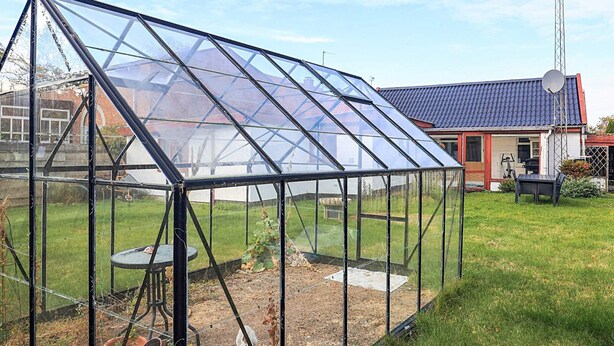 Una estructura de invernadero con techo transparente, rodeada por un césped y una casa con tejado rojo al fondo.