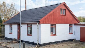 Una casa de un solo piso con techo rojo y paredes blancas, que cuenta con una chimenea, un pequeño porche y un farol.