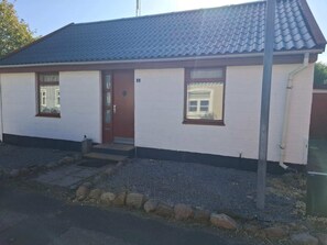 Ein kleines, einstöckiges Gebäude mit einem dunkel gefliesten Dach, weißen Wänden und einer braunen Tür. Es gibt zwei Fenster mit weißen Rahmen sowie eine kleine Veranda mit Stufen zum Eingang.