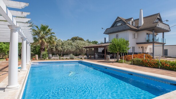 A swimming pool with a white pergola, a large house with a balcony, and a garden with flowers and palm trees.