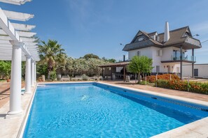 Una piscina con una pérgola blanca, una casa grande con balcón, y un jardín con flores y palmeras.